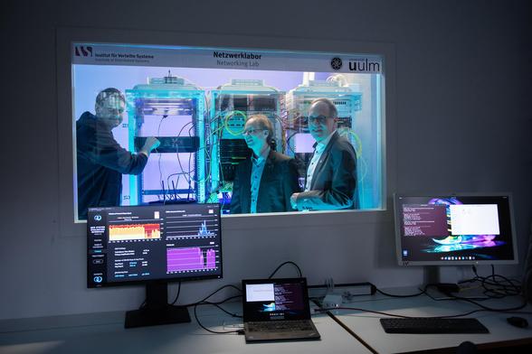 Durch eine Glasscheibe sieht man drei Männer, die vor Server-Racks stehen und im Raum davor zwei Bildschirme, die Informationen grafisch auswerten. Kevin Füchsel (links), Geschäftsführer von Quantum Optics Jena, knickt im Labor das Glasfaser-Kabel, das zu einem der Empfänger-Module führt, um einen Hackerangriff zu simulieren. Dr. Matthias Zimmermann vom DLR (Mitte) und Prof. Dr. Joachim Ankerhold (rechts) sehen zu (Foto: Elvira Eberhardt / Uni Ulm) 