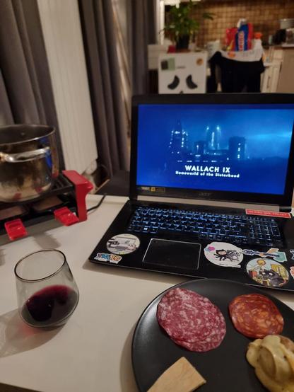 A small raclette machine with spatulas fill of cheese aroastin' and a plate with charcuterie and a potato with that good, good raclette cheese drippin' allllll over it, yeah baby. And Dune prophecy on a large laptop
