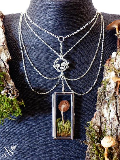 A plastron necklace with intricate cascades of silvery fine chains on a black stand in the shape of a neck. The center piece is a glass medallion with a pressed mushroom on a bed of pressed moss inside. There is a round charm above with tiny mushrooms and leaves. The overall design looks more like a harness. Alongside the stand, there are large bark pieces with real dried mushrooms on them. There’s a NS logo in one corner, with roots starting at the bottom of the N and a fern starting at the top of the S.