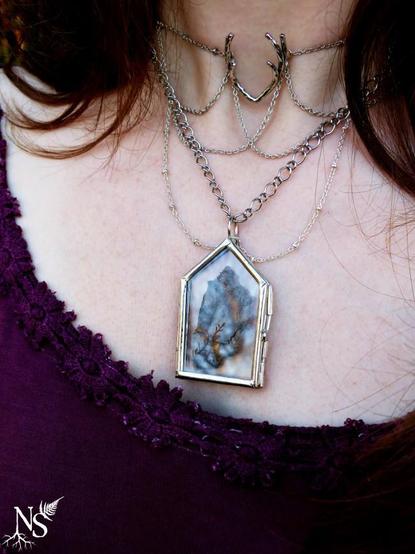 A plastron necklace with intricate cascades of silvery fine chains worn on a woman’s neck. The center piece is a glass medallion with a real piece of tree bark inside; we can see a tiny piece of red moss on it. There’s an antlers charm that falls at the crook of the neck. The fabric of her dress is a dark purple. There’s a NS logo in one corner, with roots starting at the bottom of the N and a fern starting at the top of the S.
