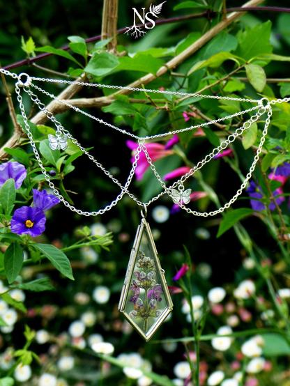 A plastron necklace with intricate cascades of silvery fine chains held tight in flowery bushes. The center piece is a glass medallion with a pressed purple flower inside (dead-nettle). There are tiny butterfly charms on the sides and they’re asymmetric on the chains, as if in flight. On the sides of the necklace, you can see that it’s really one necklace to attach that will look like multiple ones. The background is full of greenery, purple, white and fuchsia flowers. There’s a NS logo in one corner, with roots starting at the bottom of the N and a fern starting at the top of the S.