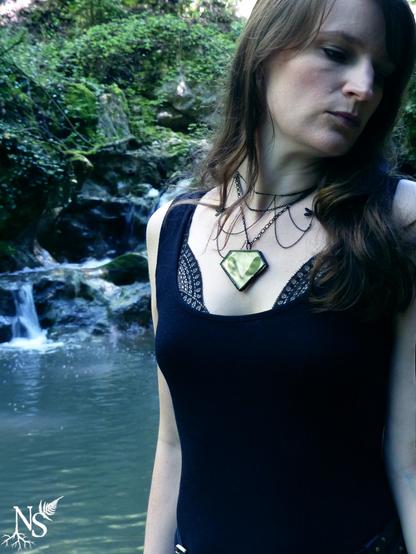 A slim woman stands before a waterfall in the forest. She’s wearing a black dress and the lace of her bra shows at the top. She has long dark blond hair and looks to her side. She’s wearing a plastron necklace with intricate cascades of black fine chains. The center piece is a glass medallion showcasing 2 real dragonfly wings. There are dragonfly charms on the sides and they’re asymmetric on the chains, as if in flight. There’s a NS logo in one corner, with roots starting at the bottom of the N and a fern starting at the top of the S.