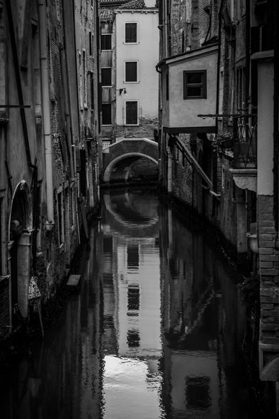 Fotografía en blanco y negro donde se ve un canal estrecho veneciano con reflejos en el agua de los edificios.