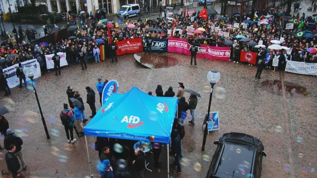 Ein AFD Wahlstand auf einem Marktplatz am unteren Bildrand, mit ein paar Menschen  die am Wahlstand stehen. 
Im weiteren, großen Kreis drumherum stehen tausend Menschen zum Gegenprotest mit Bannern und Flaggen gegen Nazis.