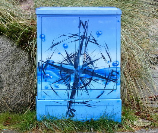 Blauer Stromkasten mit einer ebenfalls blauen Windrose bemalt die vor einer mit Gras bewachsenen Steinmauer steht