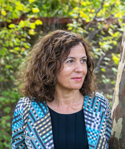 A photo of Vali happily looking off into the distance, wearing a blue diagonal check pattern blazer and a navy blue blouse.