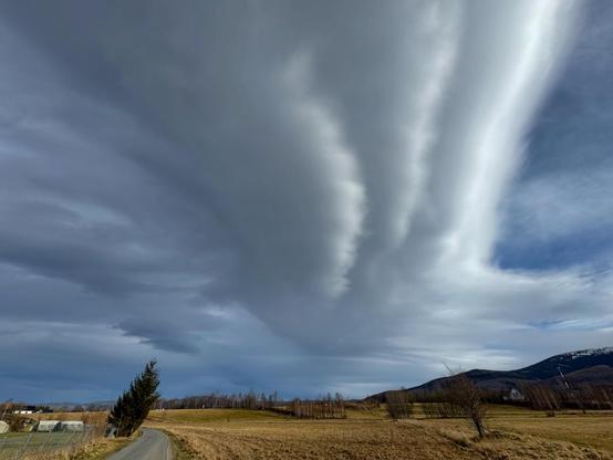 The image shows a dramatic sky with thick, elongated cloud formations stretching across the scene. The landscape includes an open field with a road, a few trees, and hills in the background. The clouds appear to form bands or streaks, creating a striking visual effect.