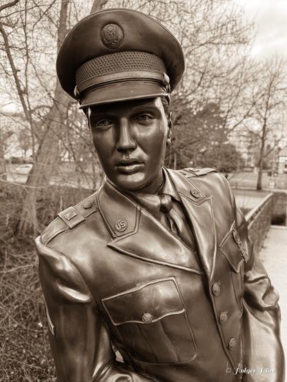 Sepia-getönte Nahaufnahme der Elvis-Bronzestatue in Bad Nauheim, Deutschland. Die Statue zeigt Elvis Presley in seiner US-Militäruniform mit ausdrucksstarkem Blick. Die warme Sepia-Farbgebung verleiht dem Bild eine nostalgische und zeitlose Atmosphäre, während die detaillierte Bronzeoberläche das Licht eindrucksvoll reflektiert.
Im Hintergrund sind kahle Bäume und eine Brücke zu sehen, die die Szene harmonisch einrahmen.
