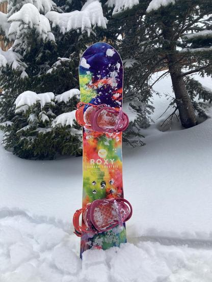 Een kleurrijk snowboard rechtop in de sneeuw, voor een tweetal besneeuwde bomen.