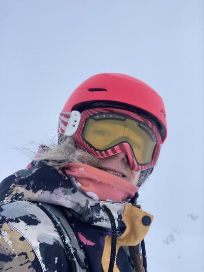 Een snowboarder met een roze helm en roze bril kijkt in de lens, de achtergrond is wit, haar haar is bevroren. Ze lacht.