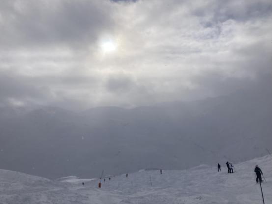 Een witte skipiste met wat personen. Op de achtergrond in de mist een silhouet van de achterliggende bergen.