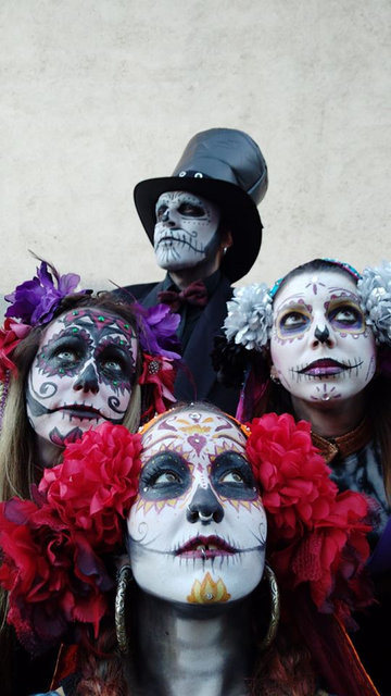 Fotografía de las caras de cuatro personas colocadas en forma de rombo. Las cuatro están maquilladas como catrinas, las calaveras mexicanas del Día de los Muertos. La cara de abajo en el centro, y las dos que están encima una a cada lado, son mujeres que además del maquillaje, llevan flores en el pelo. Arriba en el centro es un hombre con un gran sombrero de copa negro.