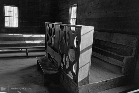 A black-and-white photo of a rustic wooden church pulpit with a divider featuring circular cutouts, lit by sunlight streaming through nearby windows.