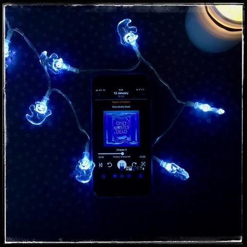 An iPhone with the cover of Only Mostly Dead (Afterlife Incorporated #1) by Alli Temple, narrated by Emily Lawrence, on.a dark background. A string of ghost fairy lights surrounds the phone and a lit candle occupies the upper right corner.
