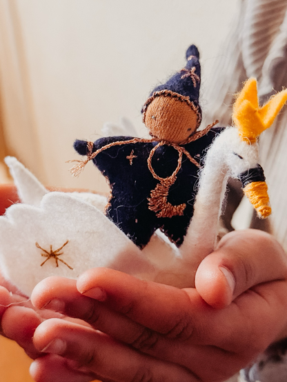 This image shows a handmade starchild or magical fairy riding a felt swan. The fairy wears a deep blue outfit with golden embroidery and a pointed hat. Its head is made of tricot fabric filled with wool. The white felt swan has delicate golden stitching and a bright yellow beak and crest. The creation, held gently in a child’s hands, exudes a whimsical, fairytale-like charm.