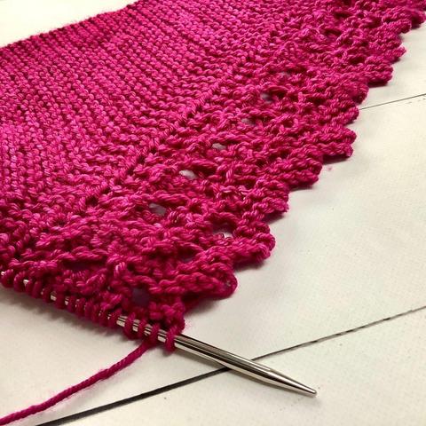 Angled view of a knitting project, featuring garter stitch and lace on a circular needle. It's shown on a white plank background.
