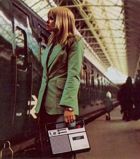 Woman in a teal jacket at a train station holding a portable cassette player.