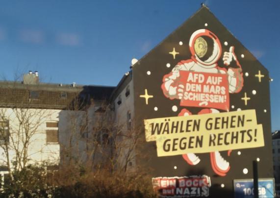 Wandgemälde in Düsseldorf-Flingern neben der Zugstrecke nach Wuppertal. Aufschrift: "Wählen gehen – gegen rechts!" Ein comichaft gezeichneter Astronautenanzug hält ein Schild "AFD auf den Mars schiessen" und reckt den linken Daumen in die Höhe. Signatur: "Kein Bock auf Nazis".