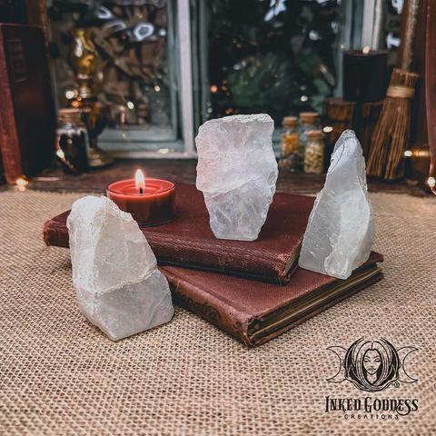 Three specimens of the "Snow Quartz Scrying Standing Stone" product are arranged around a tealight candle and old books. The crystals are about as high as they are wide with a flat bottom so that they stand upright. Presented by Inked Goddess Creations.
