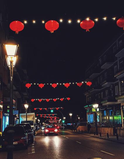 Imagen nocturna de una calle en la que se ven edificios de tres plantas de viviendas con negocios iluminados en la base. En el centro una carretera por la que circula un coche rojo de frente y sobre la que hay sucesivas hileras de farolillos rojos y redondos colgados entre los edificios, iluminando la calle. A la izquierda, coches aparcados y una farola.