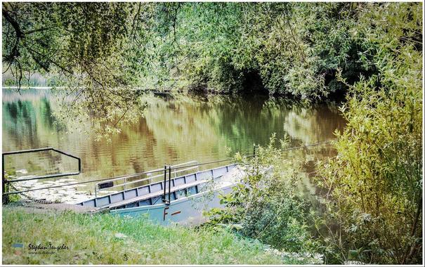 Ein idyllischer Flussabschnitt, umgeben von dichtem, grünem Laub. Im Vordergrund liegt ein kleines, metallisches Boot am Ufer, teils verdeckt von Pflanzen. Die ruhige Wasseroberfläche reflektiert die Bäume.