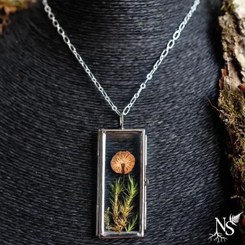 A necklace on a black jewelry display. On the side, you can make out a large piece of bark. The necklace consists of a simple chain and a glass pendant that contains a real mushroom pressed on a bed of pressed moss.
There’s a NS logo in one corner, with roots starting at the bottom of the N and a fern starting at the top of the S.
