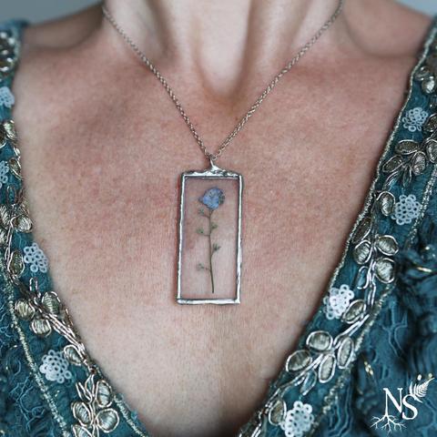 A neckline with a blue dress with silvery shiny lace and a necklace. The necklace is a simple chain and the pendant is made of stained glass and showcases a real pressed forget-me-not flower.
There’s a NS logo in one corner, with roots starting at the bottom of the N and a fern starting at the top of the S.
