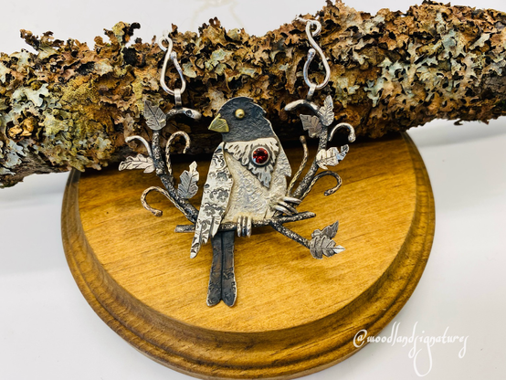 Jewelry piece featuring a silver backyard bird, a rose breasted grosbeak that looks to the left- which are known for having thick strong beaks for breaking nuts. In the center of the birds chest, features a red round garnet stone. The piece is layered to give a feel of a children’s pop up book, and around the bird perched on a branch are different leaves and tendrils of vines created from gold fill and silver. The feet of the bird are created from half circles of wire, and are very cute. The wing of the bird features floral stamping, which is one of the artists trademarks when creating wildlife pieces. 
