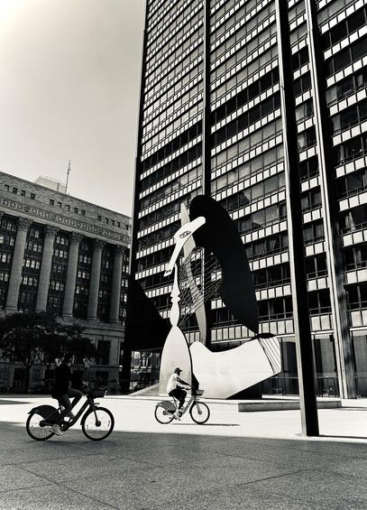 Black and white photo of two people bicycling through Daley Plaza in front of the Picasso statue. One bike is in shadow; the other is in full sun. There is a sharpe delineation between the cyclists cast by the shadow of a nearby skyscraper (out of frame).
