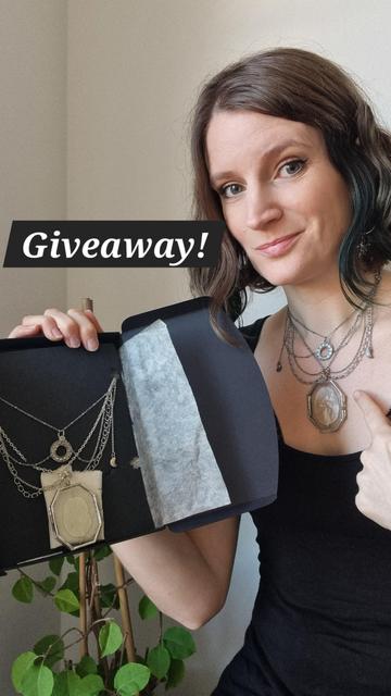 A woman points to her necklace made up of cascades of silver chains with crescent moon charms, a charm with the phases of the moon in a circle and as a central piece a glass pendant containing a silique from the Honesty (lunaria annua) with pearly reflections like the moon.
In her other hand she has an elegant black cardboard box in which a similar necklace is encased.
It says “Giveaway” in large letters because the necklace in the box is up for grabs.
