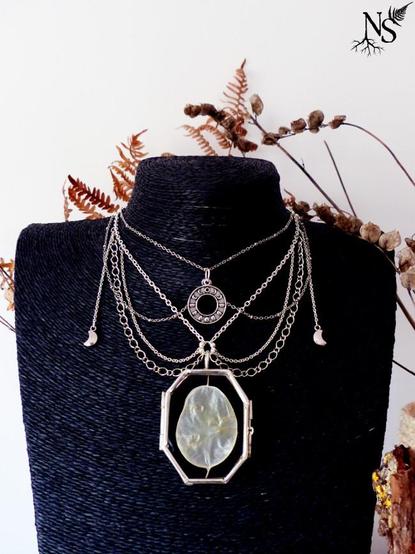 Le collier est placé sur un présentoir à colliers noir, ce qui fait très bien ressortir sa couleur argentée et le disque de lunaire. L’agencement des chaînes une fois porté est plus visible. Cela forme un collier plastron avec une partie ras de cou et certaines chaînes forment comme un col arrondi à la manière des bijoux victoriens. Quelques fleurs et fougères séchées dépassent par l’arrière.

