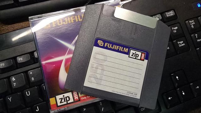 A Fujifilm 250MB Zip disk placed on top of its original packaging, which rests on a black computer keyboard. The Zip disk has a grey plastic casing with a label for writing contents and a metallic shutter at the top. The packaging underneath features a colourful futuristic design with a glowing ring and light streaks, marked as PC formatted and compatible only with 250MB Zip drives.