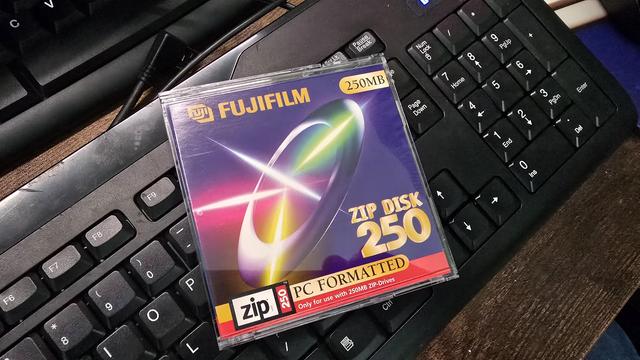 A Fujifilm Zip Disk 250MB in its original plastic case, resting on a black computer keyboard. The packaging features a colourful futuristic design with a glowing ring and streaks of light. The label indicates that the disk is PC formatted and only compatible with 250MB Zip drives.
