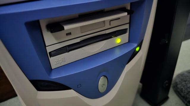 Close-up of the front panel of a vintage desktop computer tower with a blue and beige design. The panel features a Zip 250 drive with a Zip disk partially inserted, a floppy disk drive above it, and an illuminated power button. The Zip drive has a glowing green LED light, indicating activity.