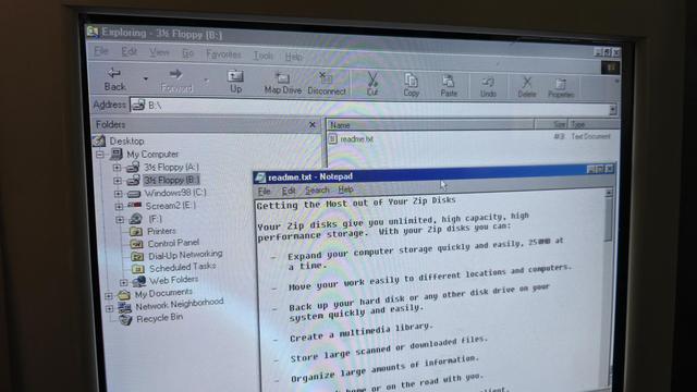 A close-up of a CRT monitor displaying a Windows 98 interface with Windows Explorer open, showing the contents of a 3.5-inch 250mb zip disk (B:). A Notepad window is open, displaying a readme.txt file about Zip disks, describing their high-capacity storage capabilities and benefits, such as expanding computer storage, backing up data, and organising files.