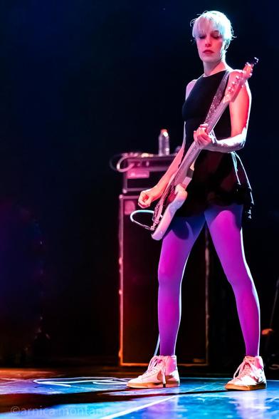 The bassist stands casually on a large stage in sneakers, bright purple tights and a black dress.