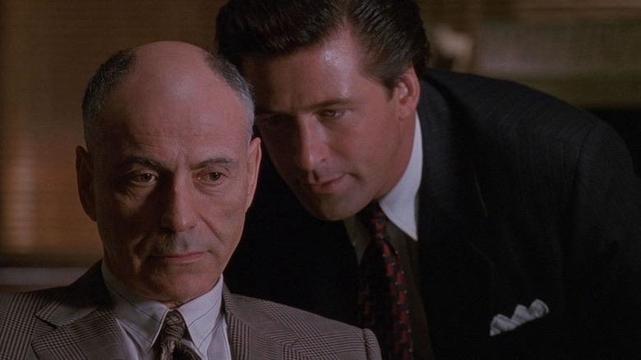 A man in a light-colored suit sits with a serious expression, while another man in a dark suit leans in closely, seemingly whispering in his ear. The background features warm lighting, suggesting a tense or intimate conversation.