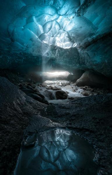 Inside an ice cave, looking towards the misty entrance. Light is coming from the entrance and a moulin above, along with cascades of water. In the foreground is a large boulder with a puddle on it, reflecting the ceiling of the ice cave.