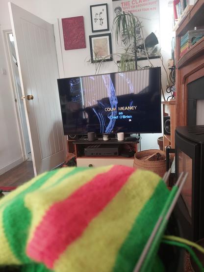 In the foreground is some bright yellow, green and orange striped knitting, and in the background is a large TV showing the opening credits to an episode of Star Trek DS9, featuring a shot of the station and the words 'Colm Meaney as Chief O'Brien'