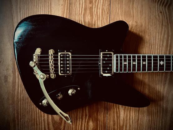 Eine schwarze E-Gitarre in „Reverse-Offset“ Form liegt auf einem Holzboden aus Kiefer. Die Gitarre hat goldene Hardware. Es ist nur der Korpus und ein kleines Stück des Halses sichtbar.