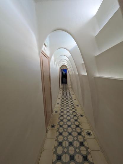 Long asymmetrical hallway opening with curved roof and arches and light slits on the right covered with white plaster and a tiled floor. 