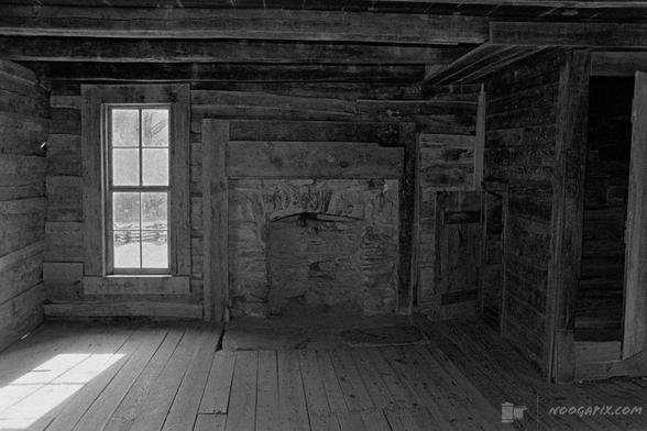 Black-and-white photo of an old wooden cabin interior with a stone fireplace, a window letting in soft light, and textured wooden walls and floors.