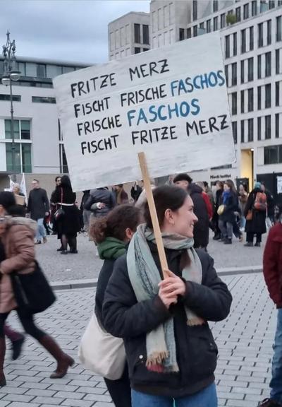 Bild einer Frau auf einer Demo mit einem Plakat in der Hand mit dem Text: "
Fritze Merz fischt frische Faschos, Frische Faschos fischt Fritze Merz"