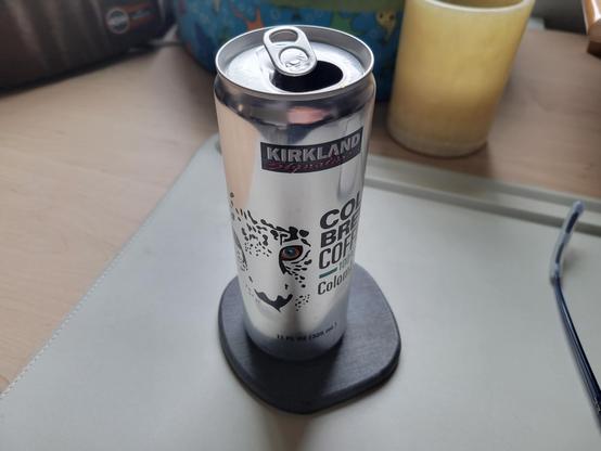 Can of Kirkland Signature Colombian Cold Brew Coffee, 11 fl oz (6-inches tall, 2-inches diameter), on a gray diatomaceous-earth coaster on an off-white desk mat on a maple desk. The can is shiny aluminum. It has the Kirkland Signature logo at top (black rectangular background, "Kirkland" in white block letters, "Signature" in red script). An illustration of the face of a leopard is at left-center, next to text "Cold Brew Coffee 100% Colombian" in black block letters (100% is in green-blue). Footer text is "11 FL OZ (325 mL)" in black font.