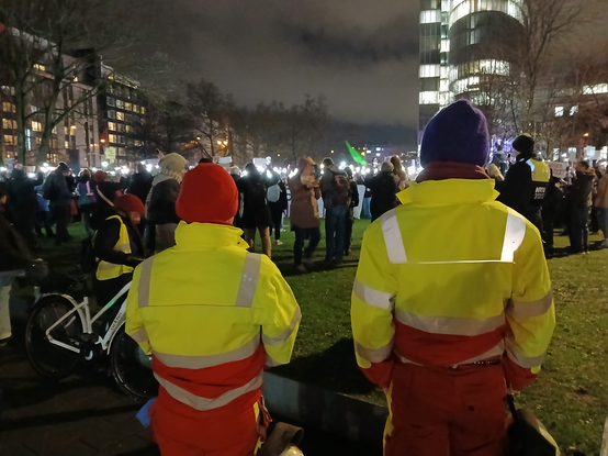 2 Demosanitäter*innen Stehen am endkundgebungsplatz, man sieht ihre Rücken und die Reflektierenden Reflektor streifen der Jacken, im Hintergrund ist die Demo Zu sehen und viele Kamera Teams die die Demo filmt, auf der Demo halten grade fast alle ihr Handy mit eingeschaltetem Blitzlicht in die Höhe und es ist ein Lichtermeer zu sehen.