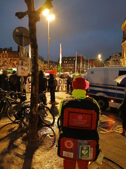 Ein Demosanitäter mit Rettungsrucksack steht vor einem Überwachungswagen der Polizei, Hinter dem Polizeiwagen ist der Anfang der Demo, welche derzeit von der Polizei Aufgehalten wird, es sind viele Demorelevante Schilder und Banner zu sehen bei denn Demonstrant*innen. 