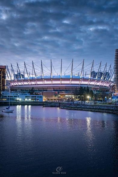A photo of a large stadium lit up with pink glowing light, shot in the evening with clouds visible in the sky and purple-reflecting water in the foreground