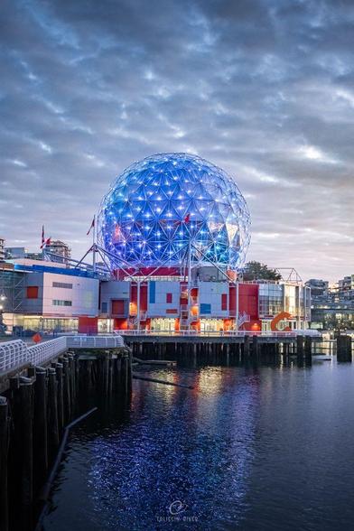 A photo of a building with a spherical top section lit up with blue lights, with dramatic grey clouds visible in the sky above, and reflections in the water below.