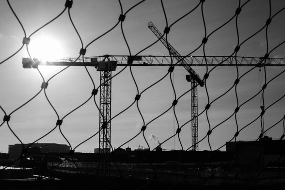 Das Bild zeigt eine urbane Szenerie in Schwarz-Weiß, die durch ein schützendes Netz im Vordergrund eingefasst wird. Die diagonalen Linien des Netzes dominieren die Komposition und erzeugen ein Gefühl von Struktur und Begrenzung, während sie die dahinterliegenden Elemente überlagern.

Hinter dem Netz sind mehrere Baukräne zu sehen, die über den Dächern der Stadt aufragen. Die Kräne symbolisieren Urbanisierung und Bauaktivität. Die Sonne steht tief am Himmel und strahlt hell, wobei sie teils hinter dem Netz und einem der Kräne verborgen ist. Dies erzeugt einen starken Kontrast und dramatische Licht- und Schatteneffekte.

Im Hintergrund sind Gebäude und die Silhouette einer Stadtlandschaft zu erkennen, ergänzt durch leichte Rauchschwaden, die auf industrielle Aktivitäten hinweisen. Der Himmel ist klar, was die Umrisse der Kräne und das Netz deutlich hervorhebt.

Das Bild vermittelt eine Kombination aus Modernität, Urbanität und Eingeschlossenheit. Die Überlagerung des Netzes erzeugt eine symbolische Trennung zwischen dem Betrachter und der Stadt, was Raum für Interpretation bietet, beispielsweise über Wachstum, Grenzen oder das Verhältnis von Mensch und urbaner Umgebung.
