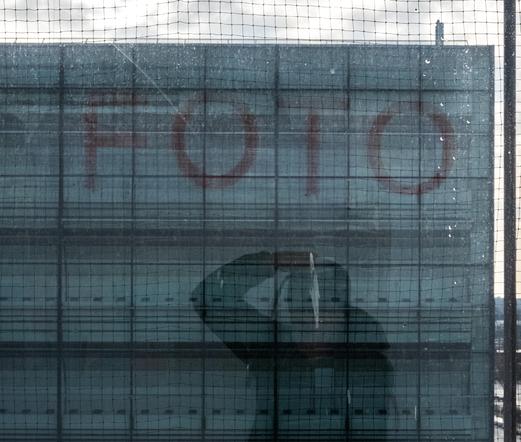Das Bild zeigt eine urbane Szene durch eine vernetzte Fensterscheibe, die den Fokus auf eine verschachtelte Komposition aus Reflexionen und der Außenwelt legt. Die Fensterscheibe ist von einem Schutzgitter oder Netz überzogen, was der Szene eine zusätzliche Ebene von Struktur und Trennung verleiht.

Im Vordergrund ist die Silhouette einer Person erkennbar, die sich möglicherweise selbst im Glas oder in der Umgebung spiegelt. Sie scheint eine Hand am Kopf oder eine Kamera vor dem Gesicht zu haben, was auf einen fotografischen Moment oder Nachdenklichkeit hindeutet. Die Reflexionen auf der Scheibe fügen eine interessante visuelle Überlagerung hinzu.

Im Hintergrund sieht man moderne Gebäude, eine breite Straße und ein Verkehrsschild. Die Szene vermittelt eine winterliche oder kühle Atmosphäre, mit kahlen Bäumen und einer leicht bewölkten Himmelslandschaft. Das Farbspiel in Blautönen unterstreicht die kühle Stimmung und betont die industriell-urbane Ästhetik.

Die Komposition wirkt ruhig, introspektiv und komplex, da sie Reflexionen, Strukturen und die Perspektive der Stadtlandschaft miteinander kombiniert. Das Bild spielt mit der Wahrnehmung von Innen und Außen, von Isolation und Verbindung.